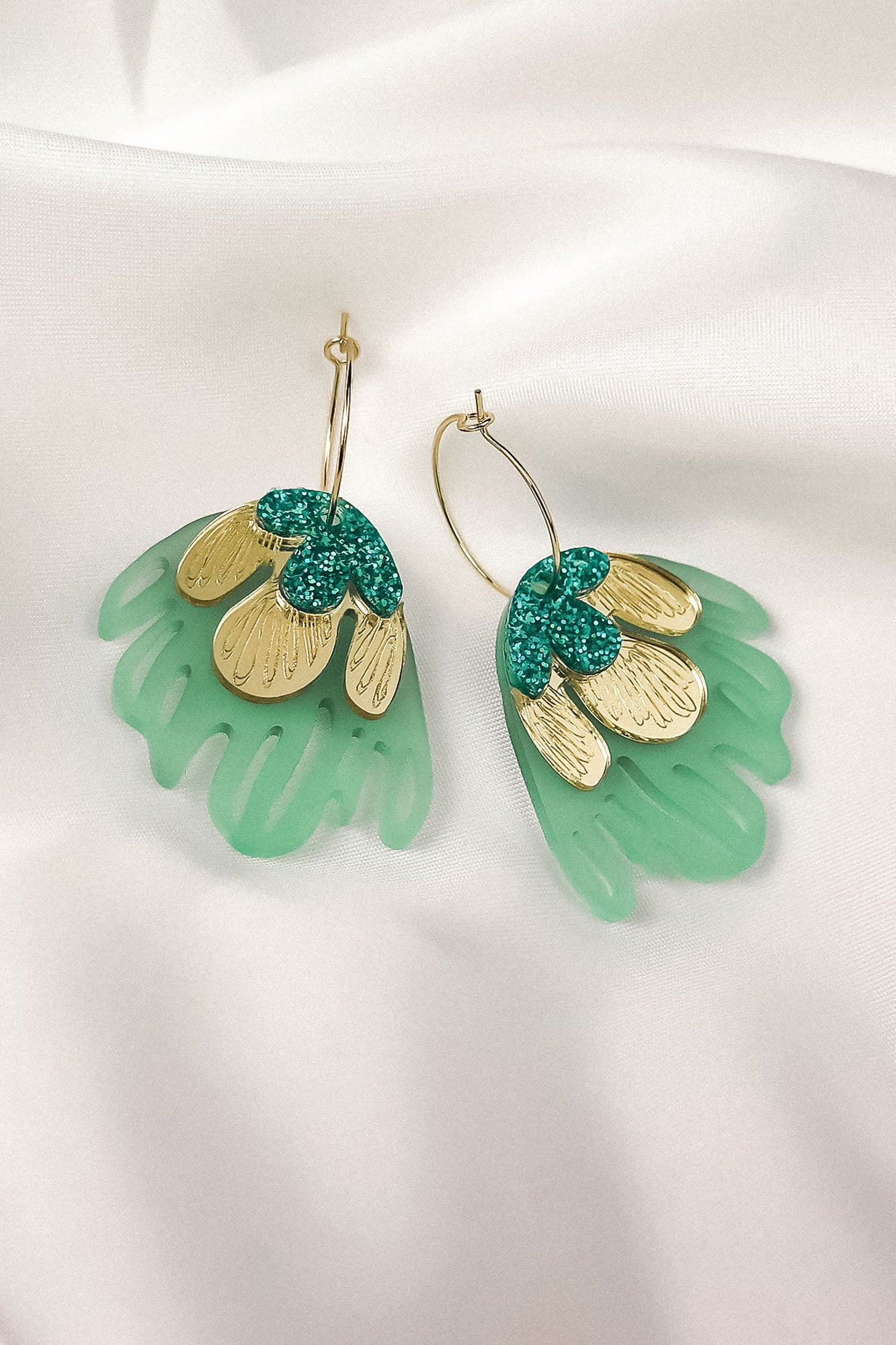 A pair of handcrafted earrings with gold metallic petals, glittery green accents, and translucent green acrylic shapes resembling leaves, displayed on white satin fabric—perfect for adding a touch of tropical elegance.