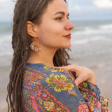 A woman with long, brown dreadlocks stands by the ocean, wearing a colorful, patterned shawl and Organic Floral Hoops accented with acrylic flowers, looking thoughtfully into the distance beneath a partly cloudy sky.