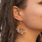 A close-up of a woman’s face with brown skin and dreadlocks, wearing vibrant Organic Floral Hoops adorned with pink, gold, and red acrylic flowers shaped like leaves or petals. The background is softly out of focus.