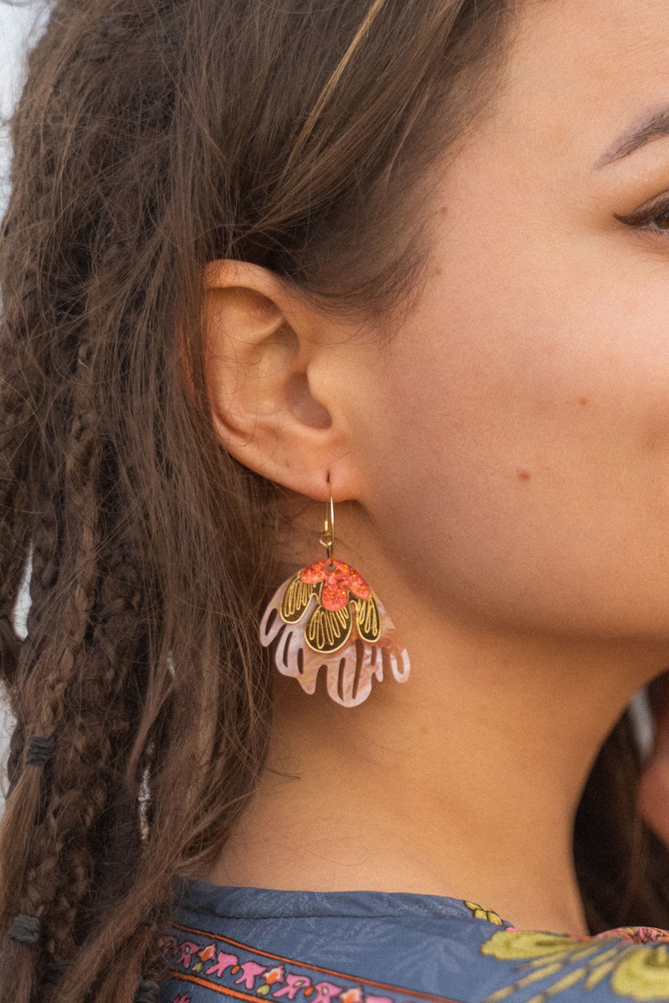 A close-up of a woman’s face with brown skin and dreadlocks, wearing vibrant Organic Floral Hoops adorned with pink, gold, and red acrylic flowers shaped like leaves or petals. The background is softly out of focus.