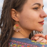 A woman with brown dreadlocks and winged eyeliner looks to the side, wearing a colorful floral-patterned garment and striking Organic Floral Hoops with pink and gold accents. She has a slight smile, her hands gently clasped.