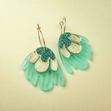 A pair of handcrafted Organic Floral Hoops with gold hoops and fan-shaped pendants. Aqua blue translucent sections and gold and teal glitter petal-like accents evoke tropical elegance against a soft, light beige background.
