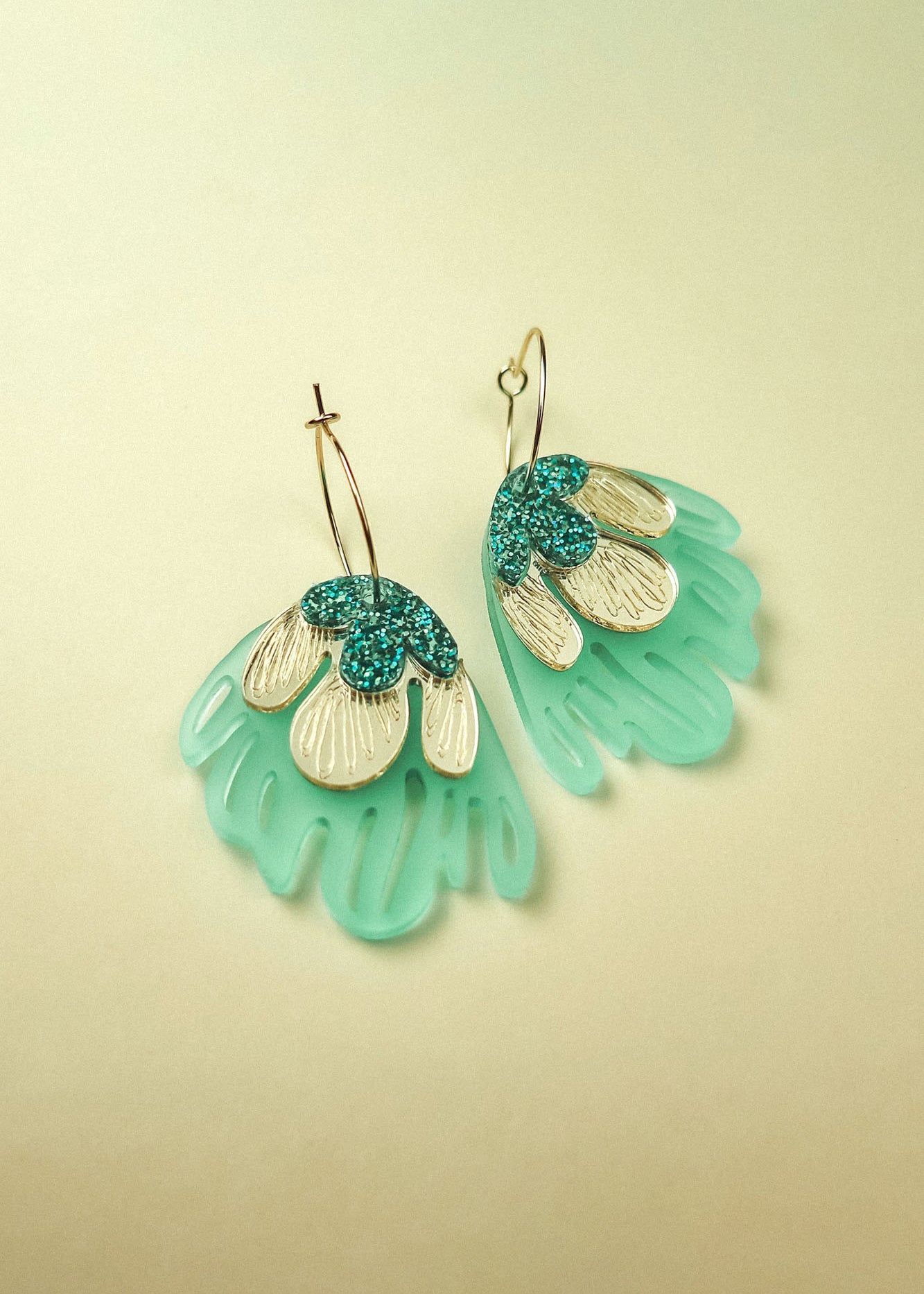 A pair of handcrafted Organic Floral Hoops with gold hoops and fan-shaped pendants. Aqua blue translucent sections and gold and teal glitter petal-like accents evoke tropical elegance against a soft, light beige background.