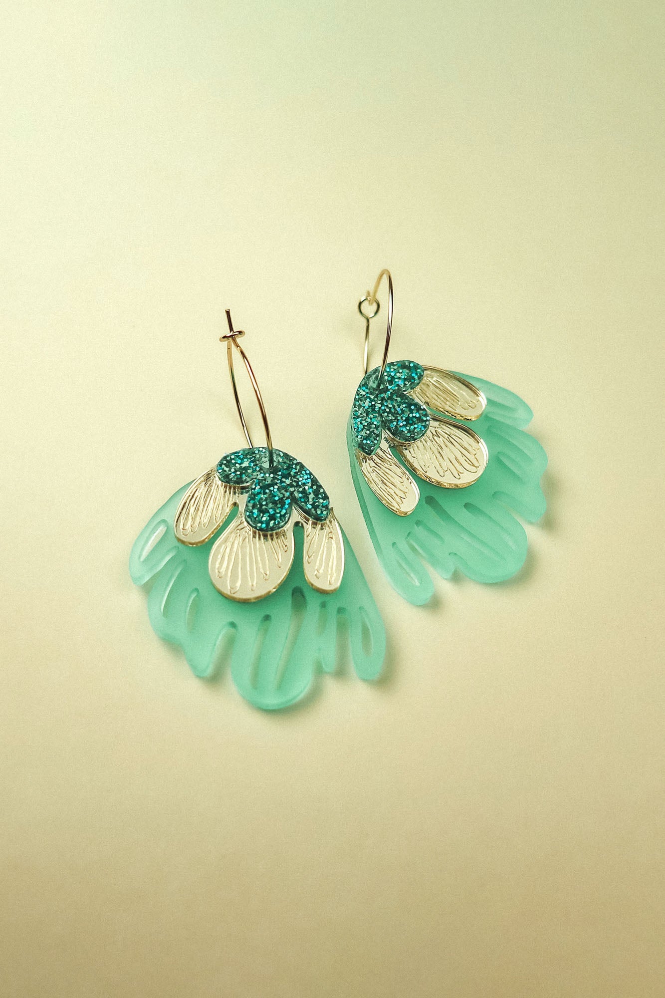 A pair of handcrafted Organic Floral Hoops with gold hoops and fan-shaped pendants. Aqua blue translucent sections and gold and teal glitter petal-like accents evoke tropical elegance against a soft, light beige background.
