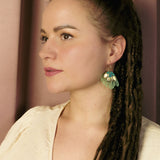 A woman with long brown dreadlocks and a cream-colored top looks to the side, wearing handcrafted, green leaf-shaped earrings that add a touch of tropical elegance. She stands in front of a mauve curtain background.