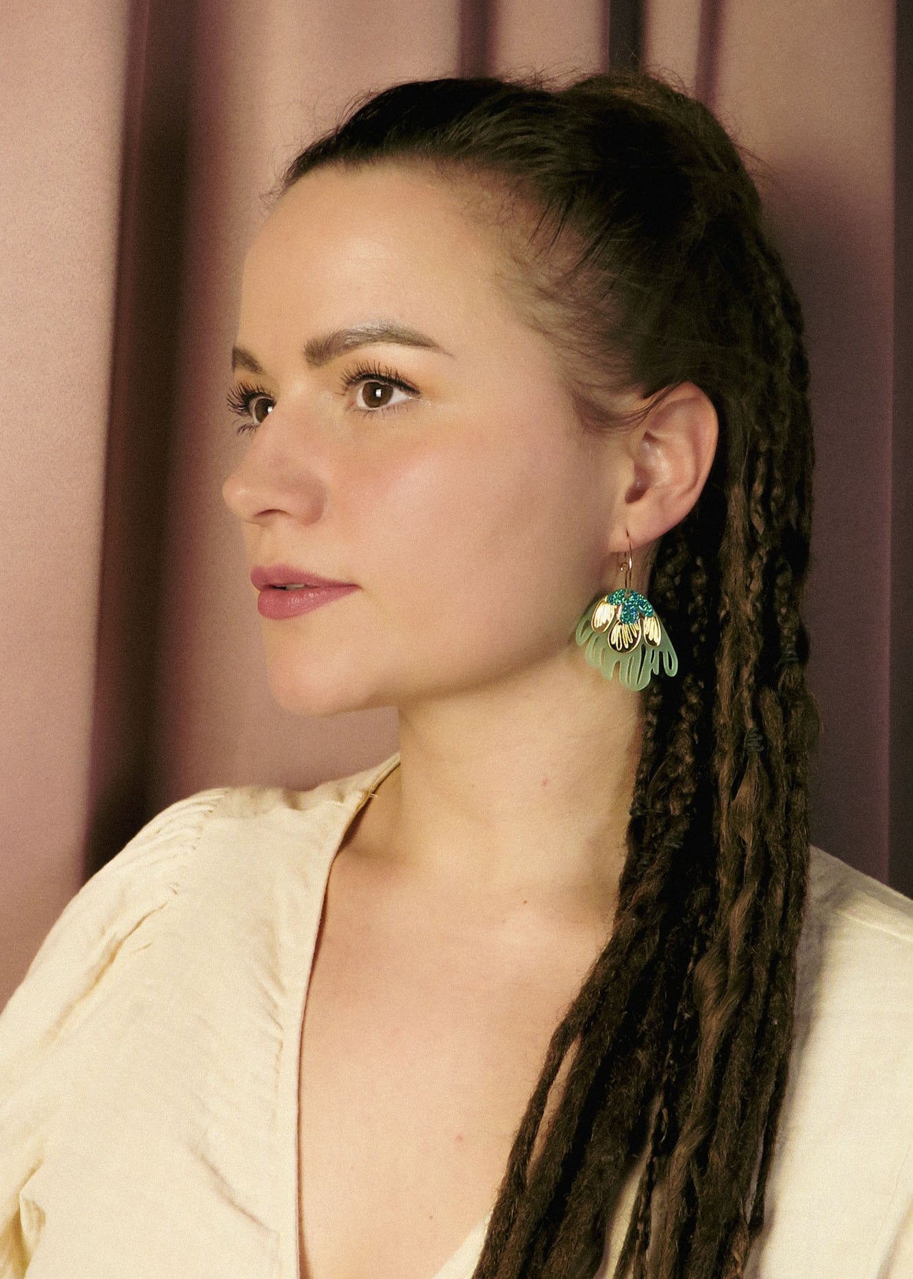 A woman with long brown dreadlocks and a cream-colored top looks to the side, wearing handcrafted, green leaf-shaped earrings that add a touch of tropical elegance. She stands in front of a mauve curtain background.