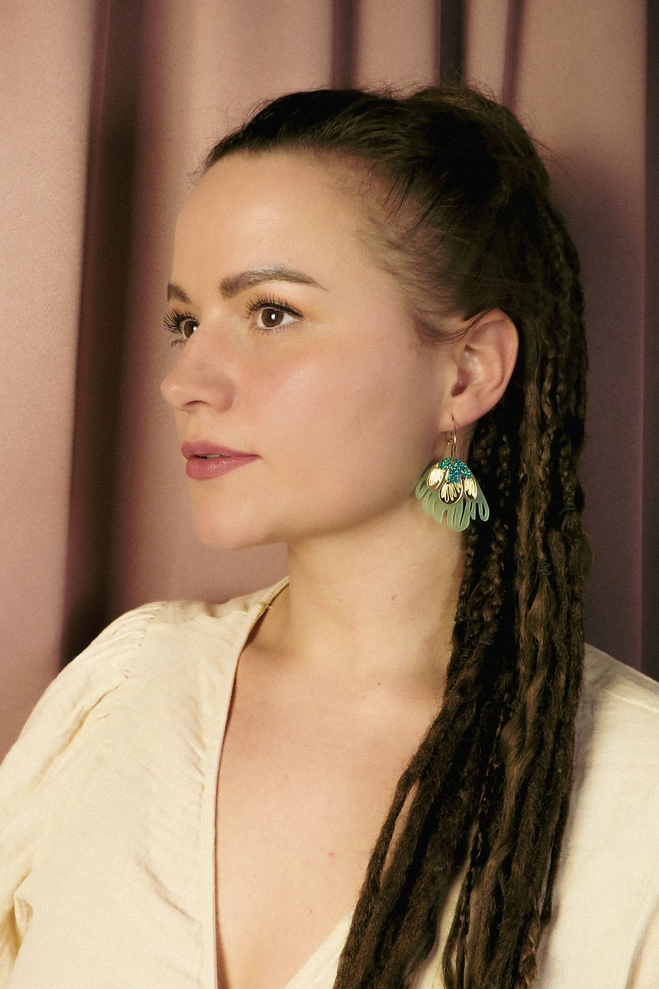 A woman with long brown dreadlocks and a cream-colored top looks to the side, wearing handcrafted, green leaf-shaped earrings that add a touch of tropical elegance. She stands in front of a mauve curtain background.