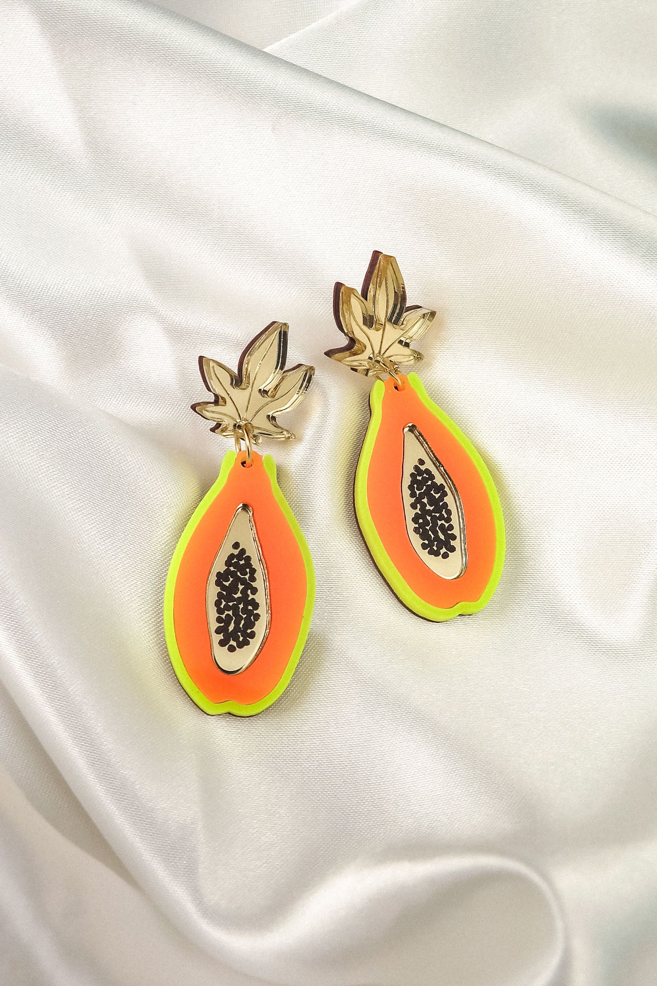 Add a playful pop to your look with these Papaya Earrings, crafted from neon acrylic glass and featuring allergy-free stainless steel hooks. The colorful halves with gold leaves and black seeds are displayed on smooth white satin fabric.