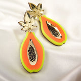 A pair of colorful Papaya Earrings shaped like papaya slices, crafted from neon acrylic glass with gold leaf tops, orange and yellow accents, black seed details, and allergy-free stainless steel posts, displayed on white satin fabric.