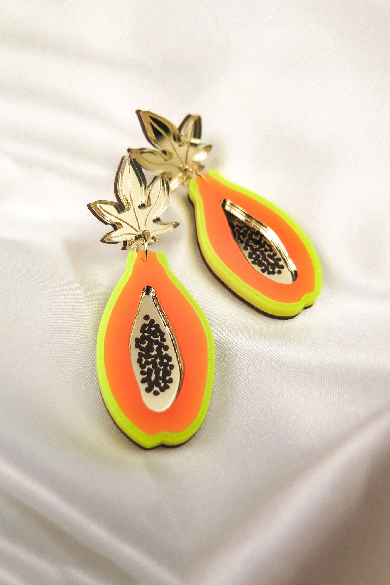 A pair of colorful Papaya Earrings shaped like papaya slices, crafted from neon acrylic glass with gold leaf tops, orange and yellow accents, black seed details, and allergy-free stainless steel posts, displayed on white satin fabric.