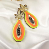 A pair of colorful Papaya Earrings shaped like papaya halves, crafted from neon acrylic glass with gold, orange, and neon green layers, featuring black seed details. Allergy-free stainless steel posts ensure comfort. Displayed on white satin with dried foliage behind.