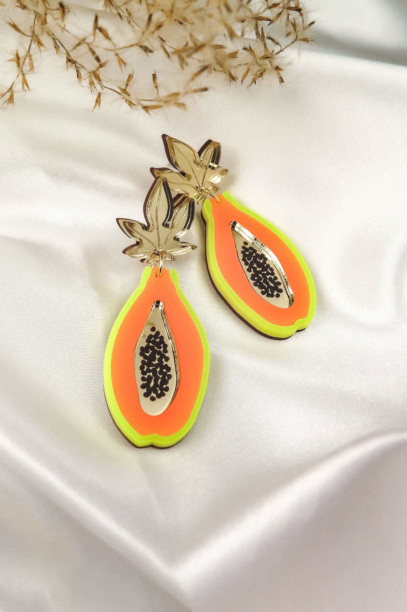 A pair of colorful Papaya Earrings shaped like papaya halves, crafted from neon acrylic glass with gold, orange, and neon green layers, featuring black seed details. Allergy-free stainless steel posts ensure comfort. Displayed on white satin with dried foliage behind.
