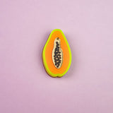 A handcrafted Papaya Pin shaped like a papaya slice features neon orange, yellow, and green layers with black seeds, set against a light purple background.