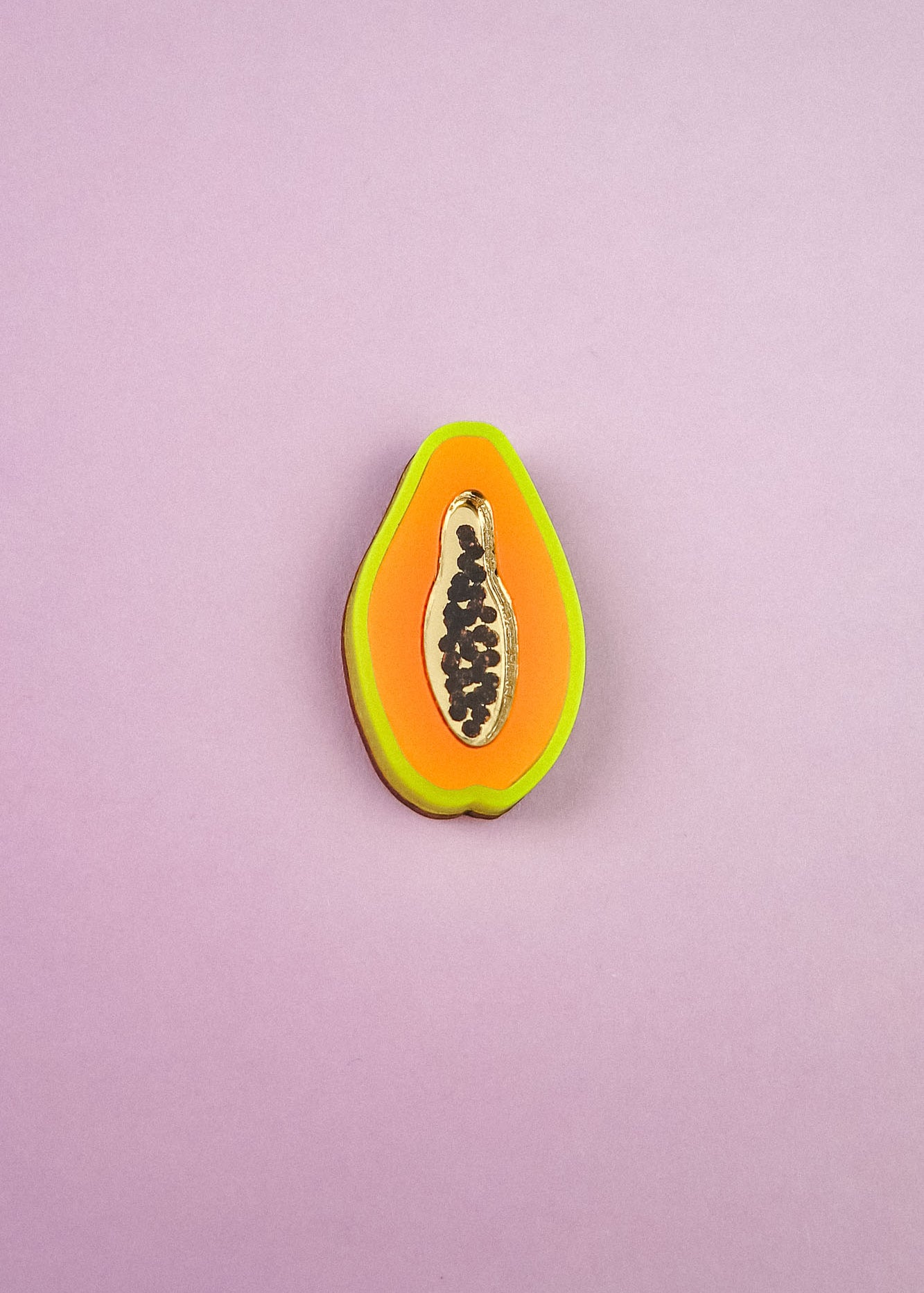A handcrafted Papaya Pin shaped like a papaya slice features neon orange, yellow, and green layers with black seeds, set against a light purple background.
