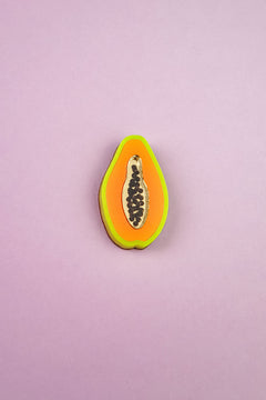 A handcrafted Papaya Pin shaped like a papaya slice features neon orange, yellow, and green layers with black seeds, set against a light purple background.