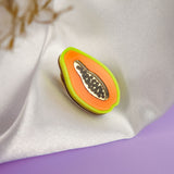 A handcrafted Papaya Pin, featuring a colorful enamel design with neon accents and black seeds, rests on white fabric against a soft lavender surface and a blurred beige plant background.