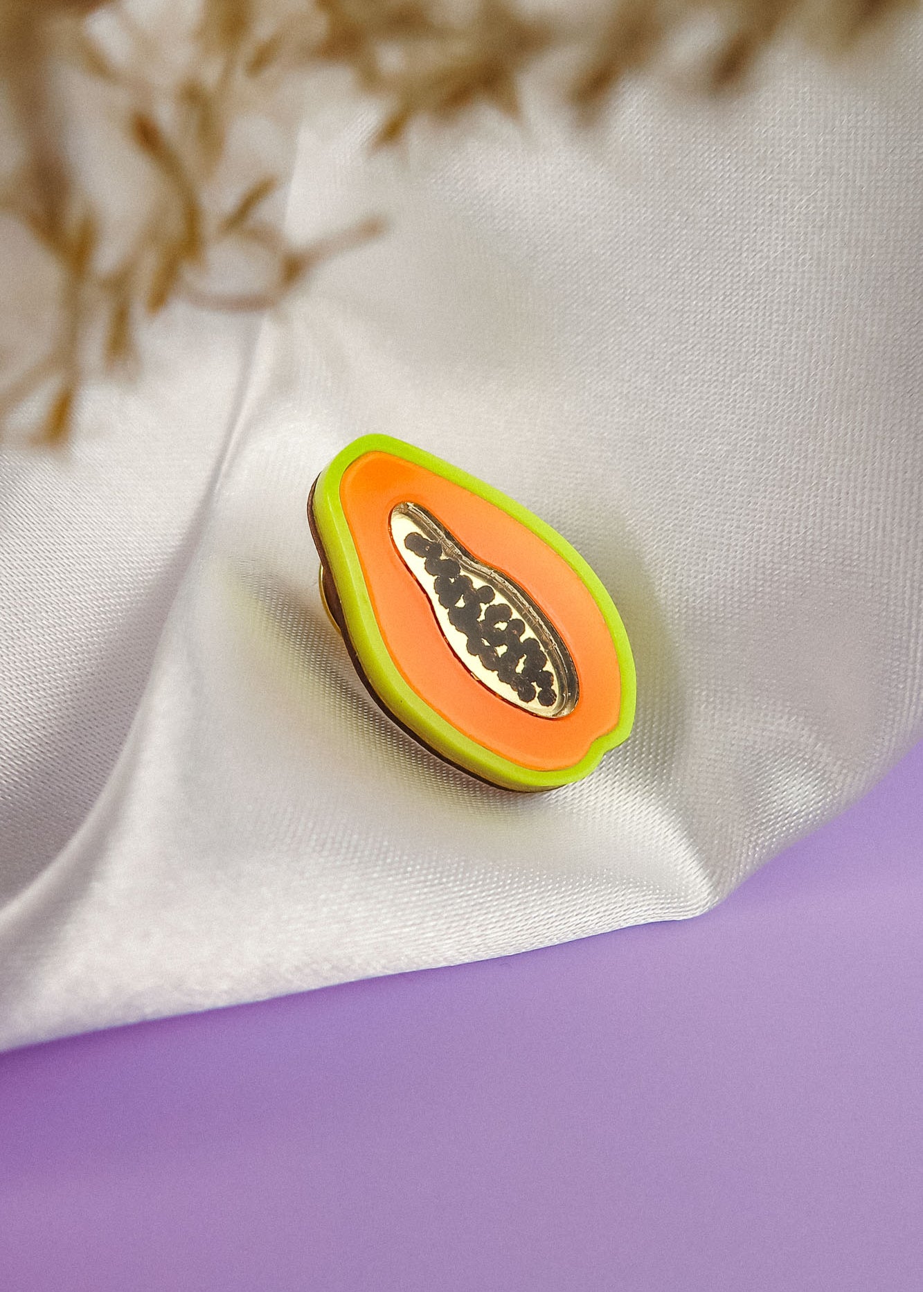 A handcrafted Papaya Pin, featuring a colorful enamel design with neon accents and black seeds, rests on white fabric against a soft lavender surface and a blurred beige plant background.
