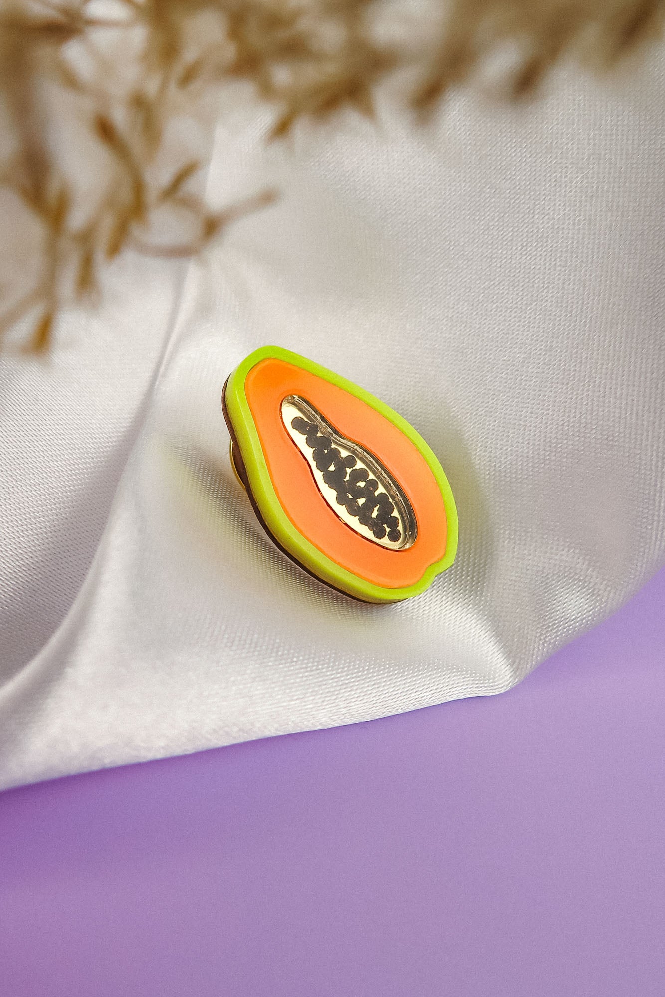 A handcrafted Papaya Pin, featuring a colorful enamel design with neon accents and black seeds, rests on white fabric against a soft lavender surface and a blurred beige plant background.