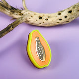 A handcrafted, colorful papaya-shaped cookie with icing details sits on a purple surface next to a dried, spotted branch, reminiscent of a neon Papaya Pin.