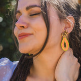 A woman with long brown hair smiles softly with her eyes closed, wearing bright Papaya Earrings made from allergy-free stainless steel and a sheer white top, standing outdoors in sunlight.