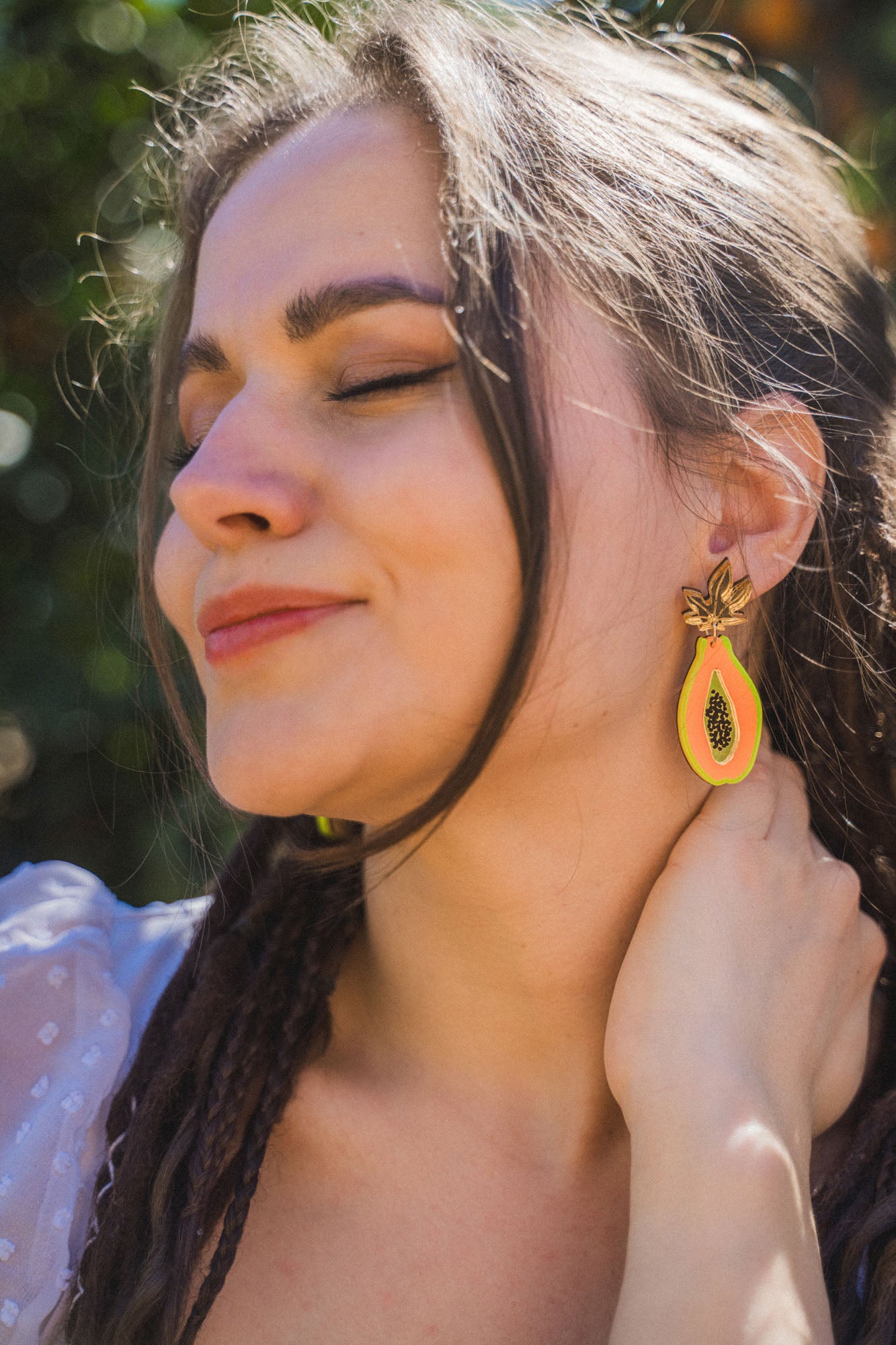 A woman with long brown hair smiles softly with her eyes closed, wearing bright Papaya Earrings made from allergy-free stainless steel and a sheer white top, standing outdoors in sunlight.
