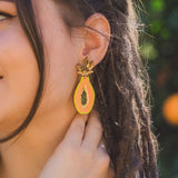 A woman with braided hair wears vibrant Papaya Earrings crafted from allergy-free stainless steel and neon acrylic glass, gently touching one as she stands before blurred greenery.