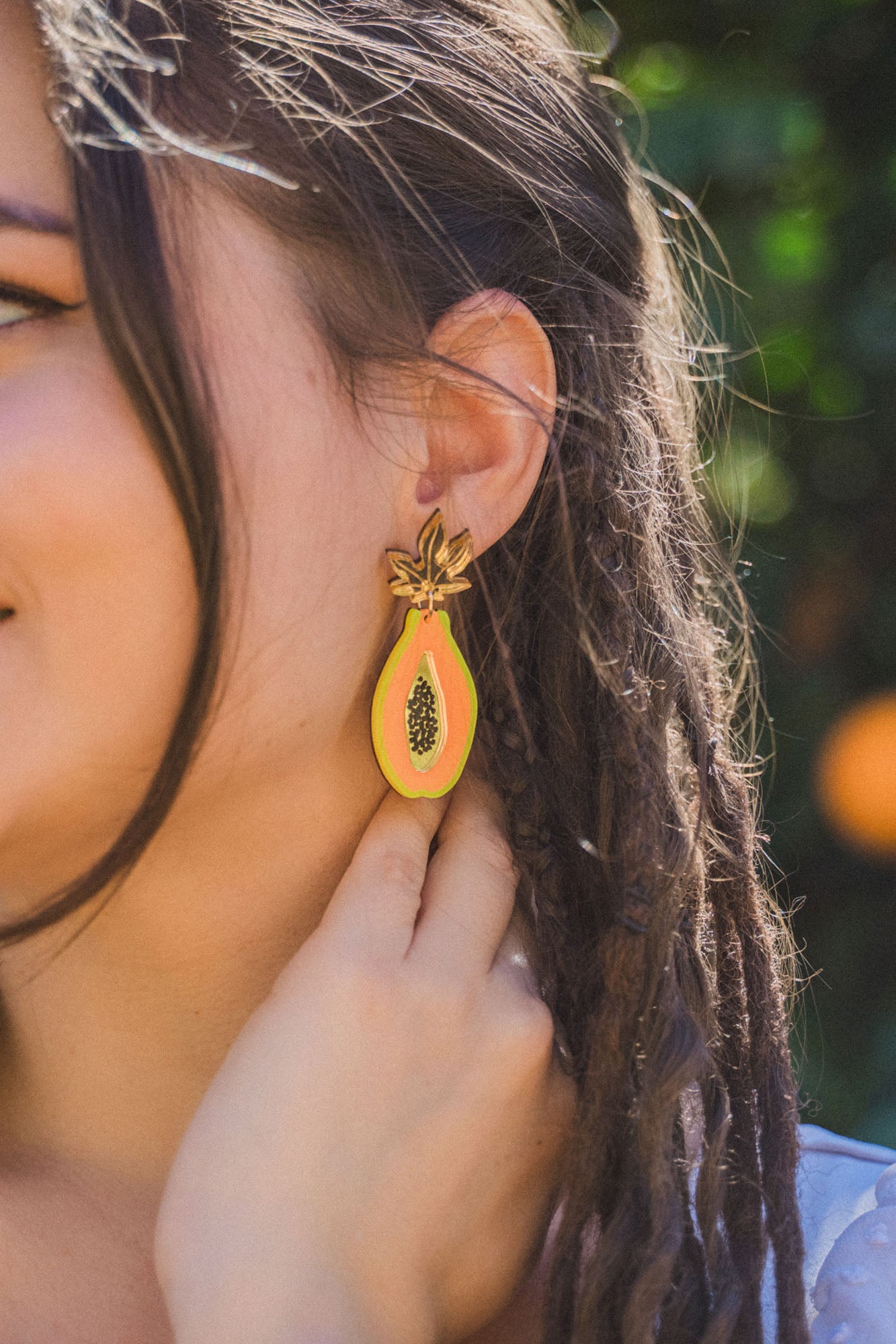 A woman with braided hair wears vibrant Papaya Earrings crafted from allergy-free stainless steel and neon acrylic glass, gently touching one as she stands before blurred greenery.