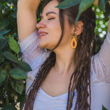 A woman with long dreadlocks and a tattoo on her arm stands among green leaves, eyes closed and smiling, wearing a white top and allergy-free stainless steel Papaya Earrings in bright orange.