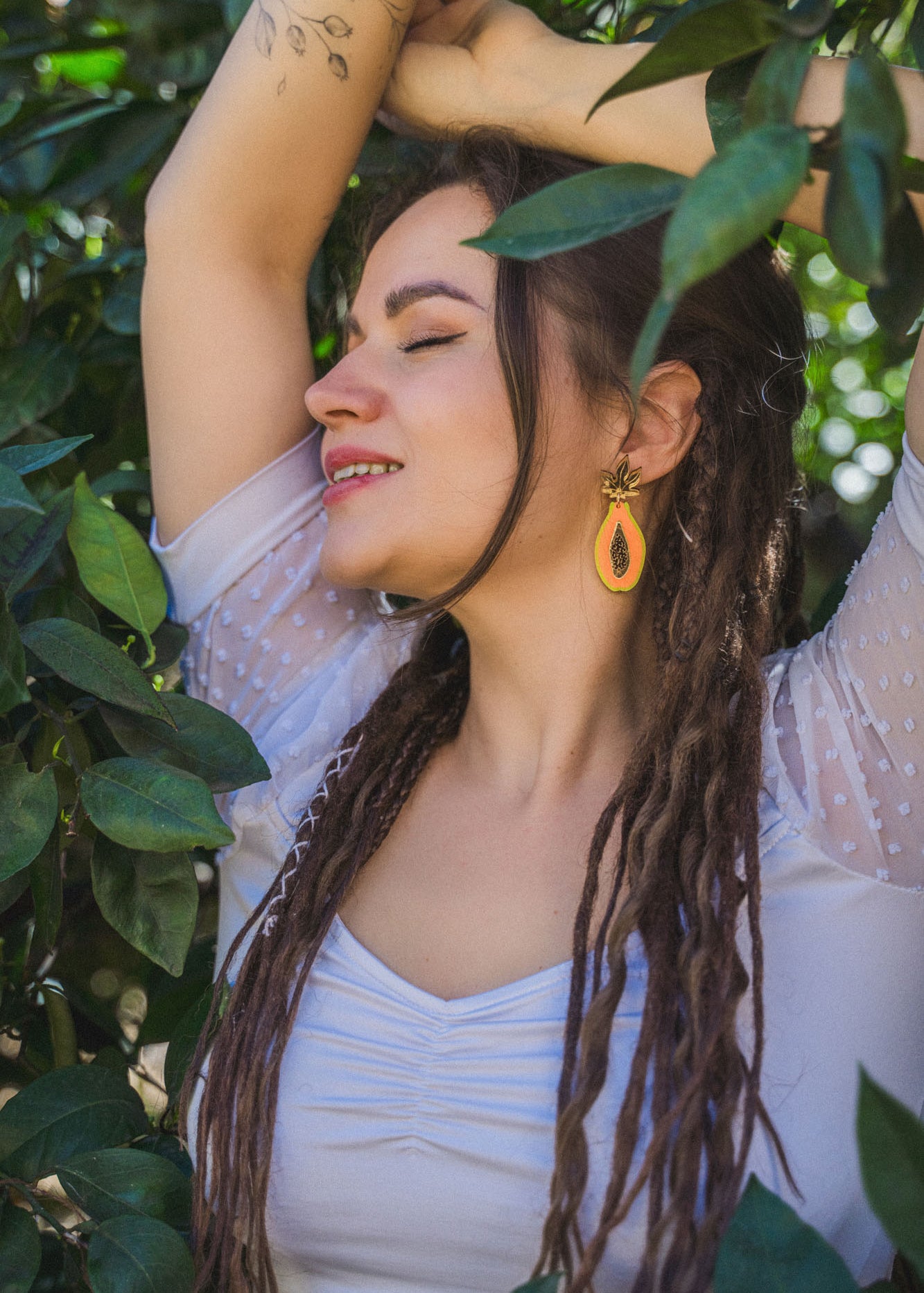 A woman with long dreadlocks and a tattoo on her arm stands among green leaves, eyes closed and smiling, wearing a white top and allergy-free stainless steel Papaya Earrings in bright orange.