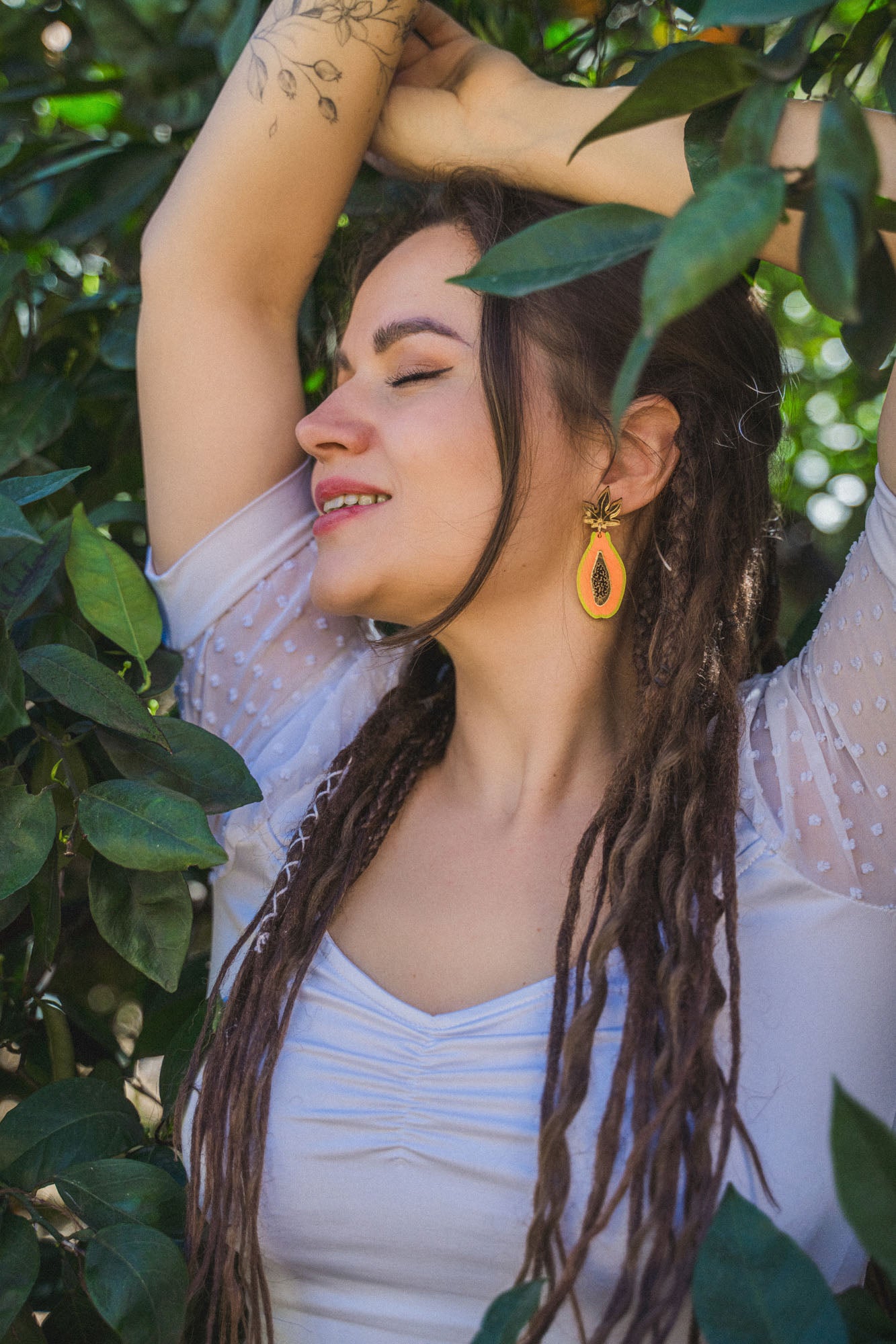 A woman with long dreadlocks and a tattoo on her arm stands among green leaves, eyes closed and smiling, wearing a white top and allergy-free stainless steel Papaya Earrings in bright orange.