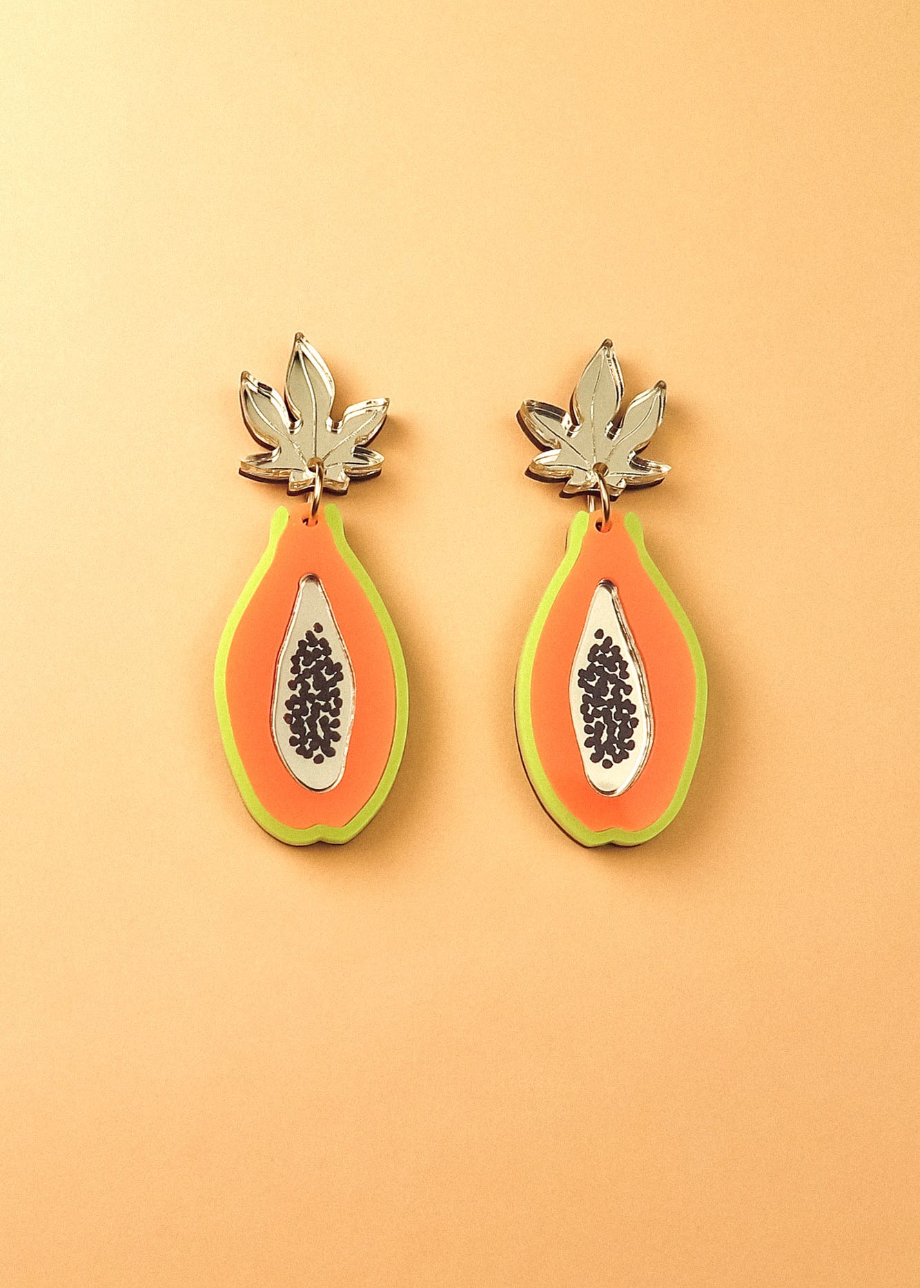 Papaya Earrings made of allergy-free stainless steel and neon acrylic glass, featuring orange with green edges, black seed details, and silver leaves, all set against a peach-colored background.