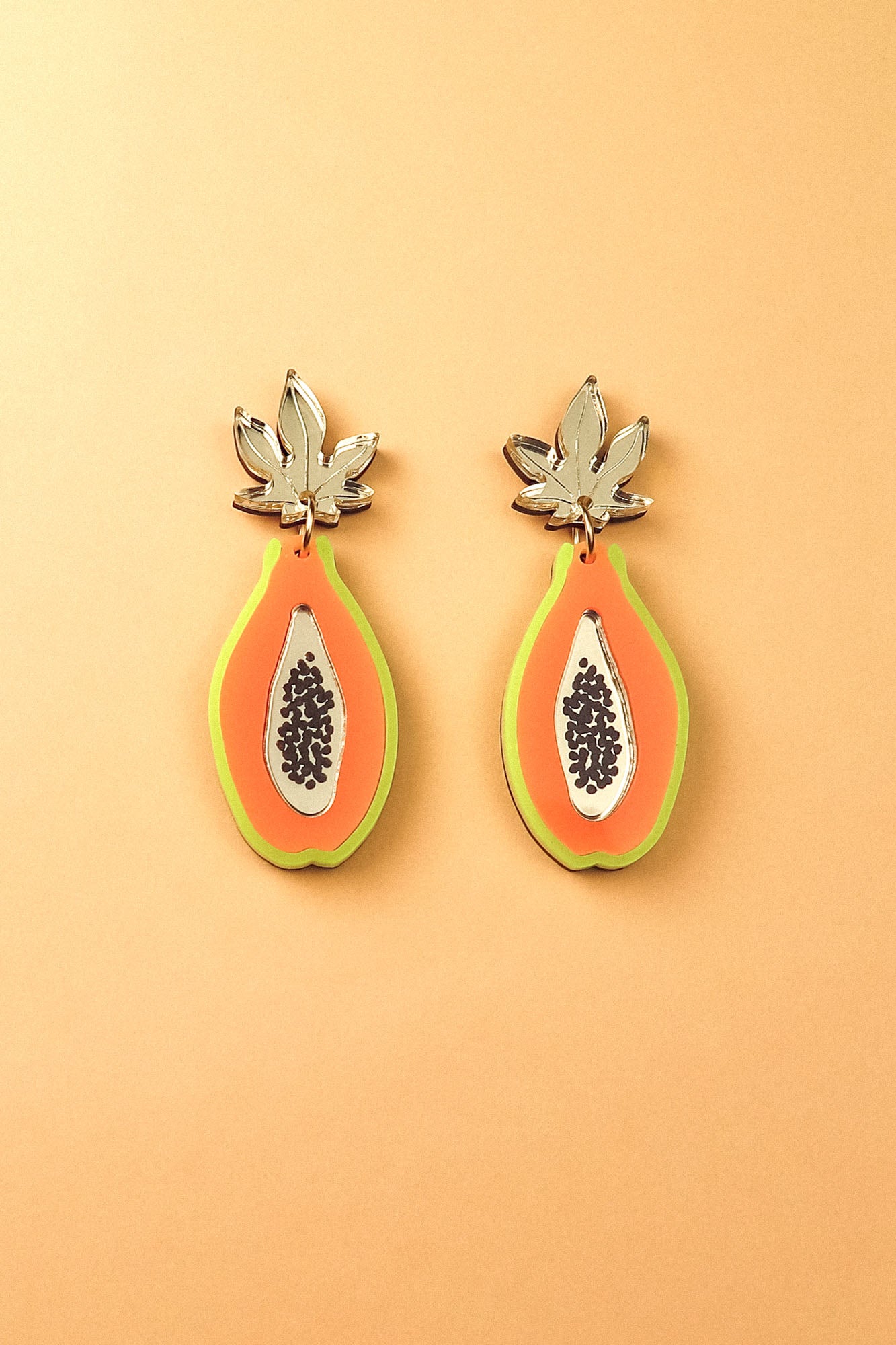 Papaya Earrings made of allergy-free stainless steel and neon acrylic glass, featuring orange with green edges, black seed details, and silver leaves, all set against a peach-colored background.