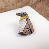 A geometric Penguin Pin accessory, crafted from wood in black, white, and yellow sections, rests on a white surface. Wispy beige plants are blurred in the background, making this brooch a charming addition to any outfit.
