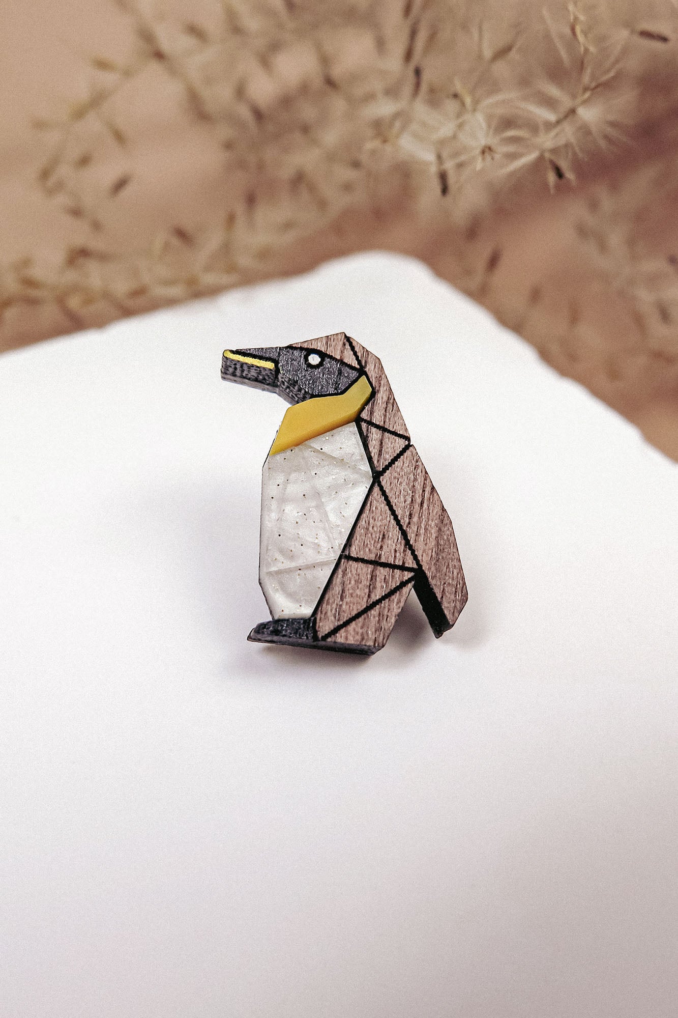 A geometric Penguin Pin accessory, crafted from wood in black, white, and yellow sections, rests on a white surface. Wispy beige plants are blurred in the background, making this brooch a charming addition to any outfit.
