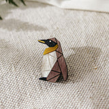 A geometric, wooden Penguin Pin with black, brown, white, and yellow sections sits on a textured beige fabric, making a charming accessory to enhance any outfit. Green fern leaves are blurred in the background.