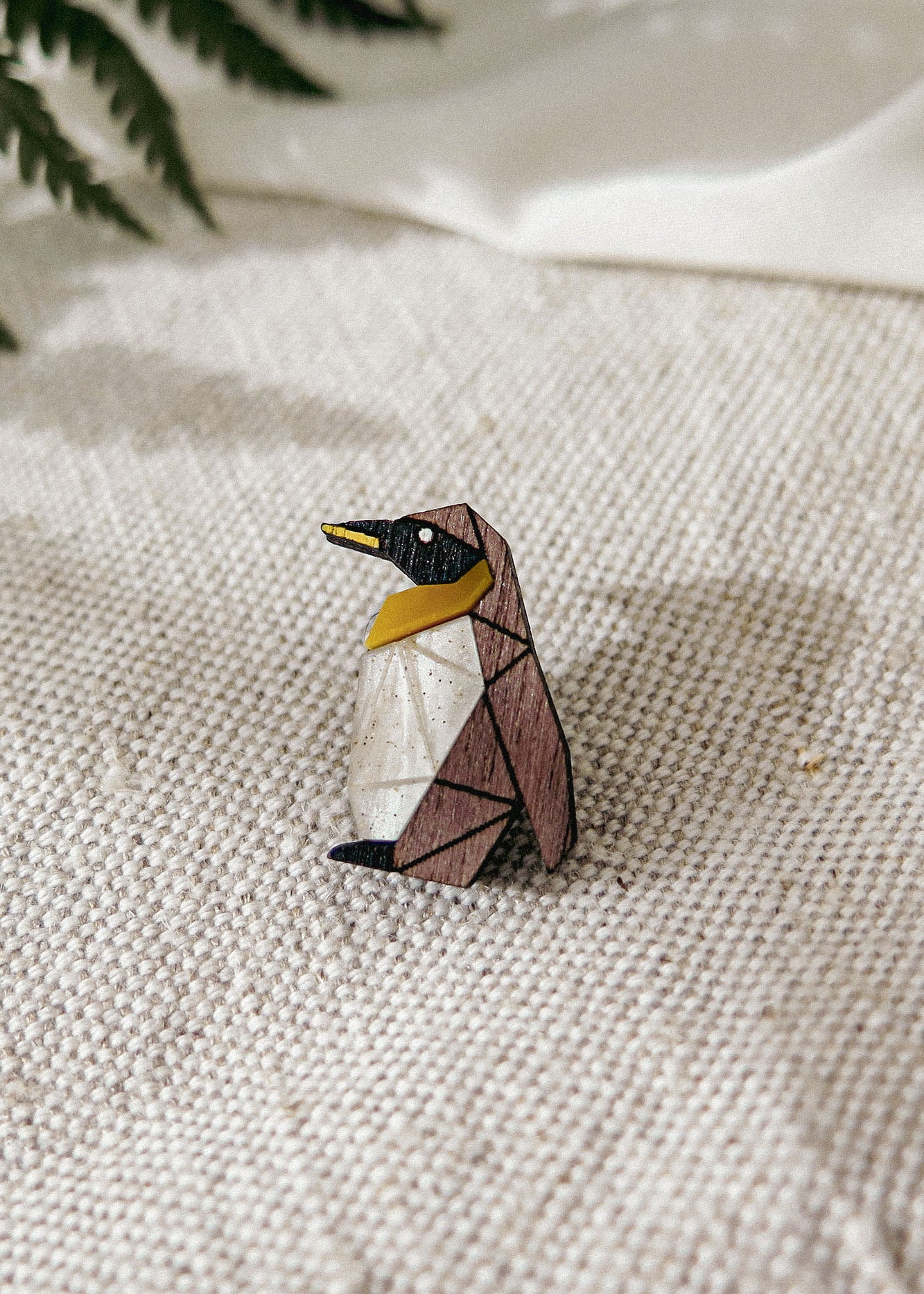 A geometric, wooden Penguin Pin with black, brown, white, and yellow sections sits on a textured beige fabric, making a charming accessory to enhance any outfit. Green fern leaves are blurred in the background.