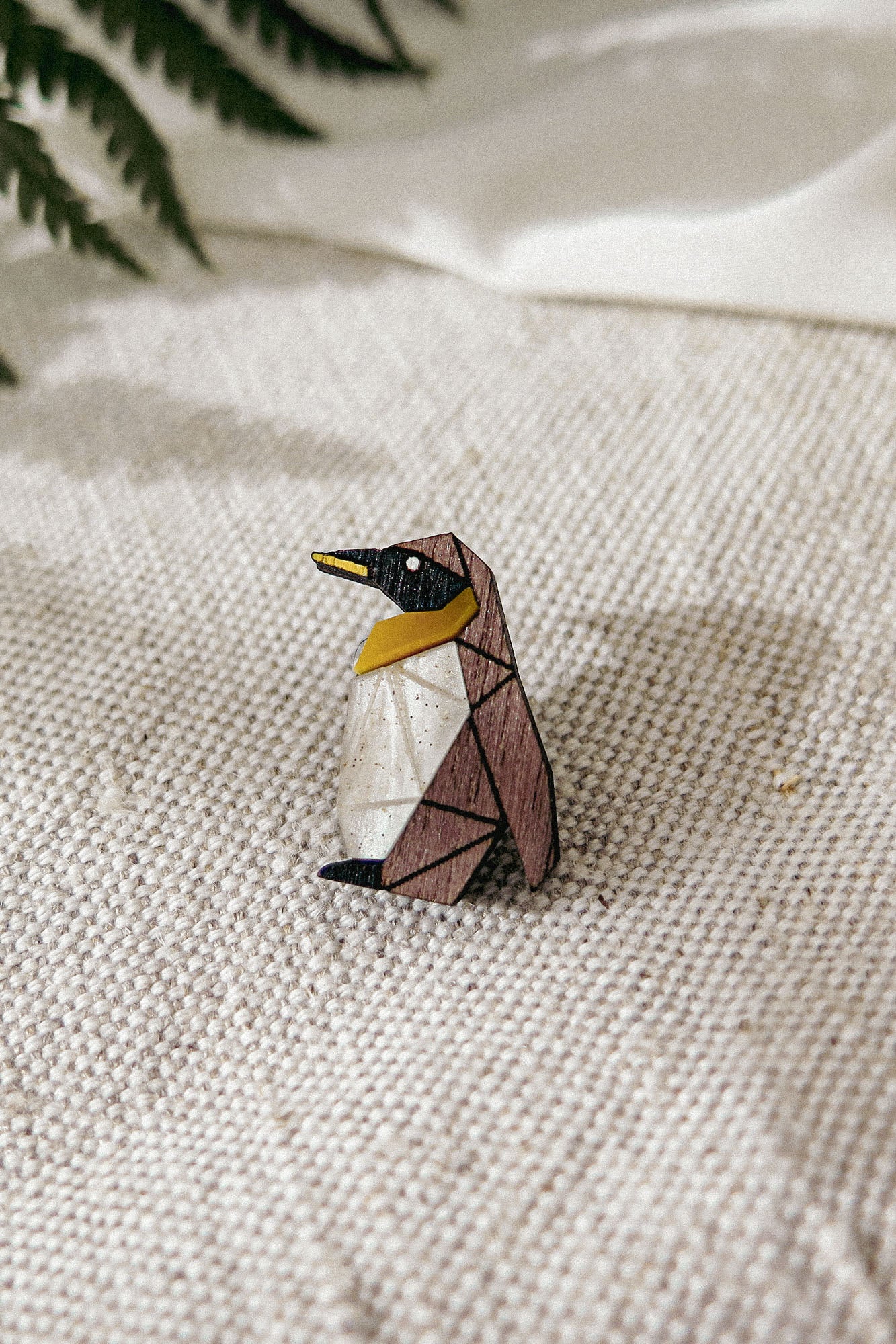 A geometric, wooden Penguin Pin with black, brown, white, and yellow sections sits on a textured beige fabric, making a charming accessory to enhance any outfit. Green fern leaves are blurred in the background.