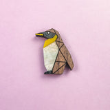 A wooden, geometric Penguin Pin with yellow, white, and black accents serves as a charming accessory, set against a light pink background.