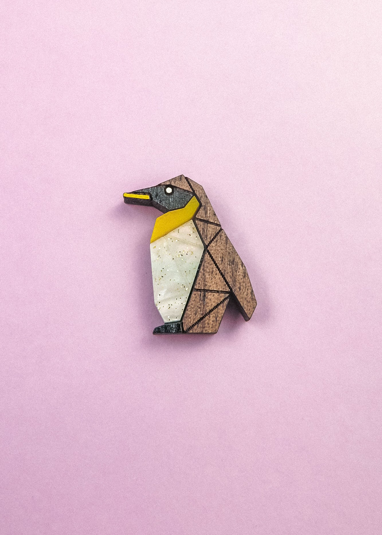 A wooden, geometric Penguin Pin with yellow, white, and black accents serves as a charming accessory, set against a light pink background.