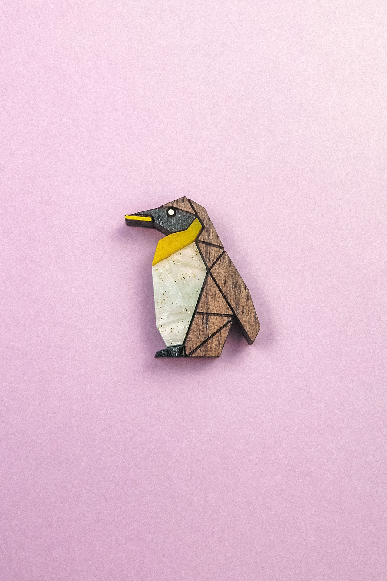 A wooden, geometric Penguin Pin with yellow, white, and black accents serves as a charming accessory, set against a light pink background.