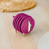 A set of round, maroon felt coasters stacked vertically on a metal holder shaped like a sheep, placed on a light wooden table near woven placemats—an ideal housewarming gift.