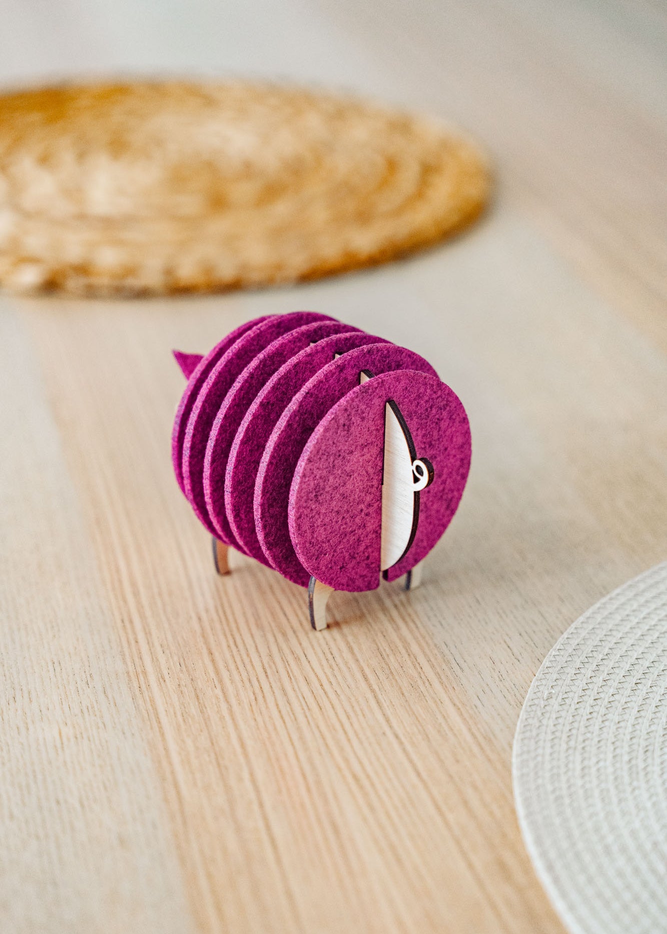 A set of round, maroon felt coasters stacked vertically on a metal holder shaped like a sheep, placed on a light wooden table near woven placemats—an ideal housewarming gift.