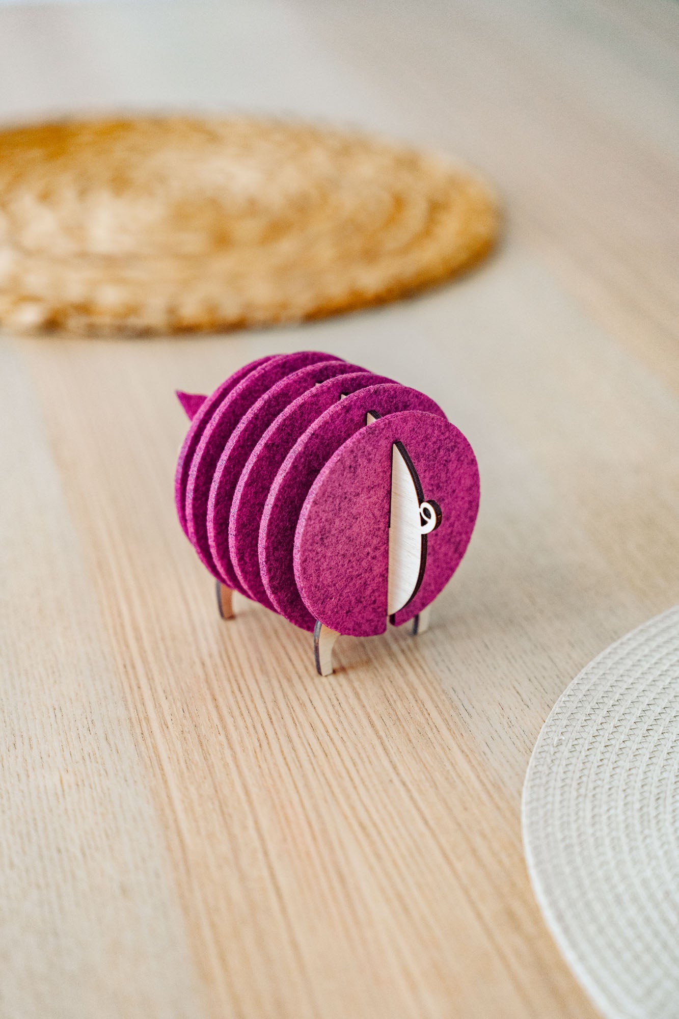 A set of round, maroon felt coasters stacked vertically on a metal holder shaped like a sheep, placed on a light wooden table near woven placemats—an ideal housewarming gift.
