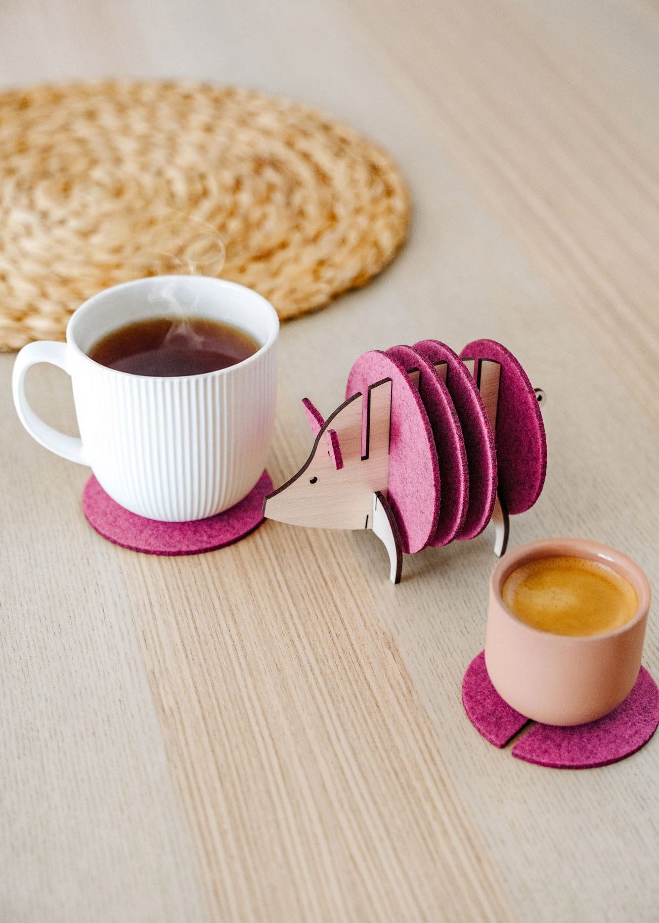 A white cup and a pink cup sit on purple felt coasters next to a wooden hedgehog-shaped coaster holder, with more felt coasters inside it, on a light wooden table—a lovely housewarming gift. A woven mat is in the background.