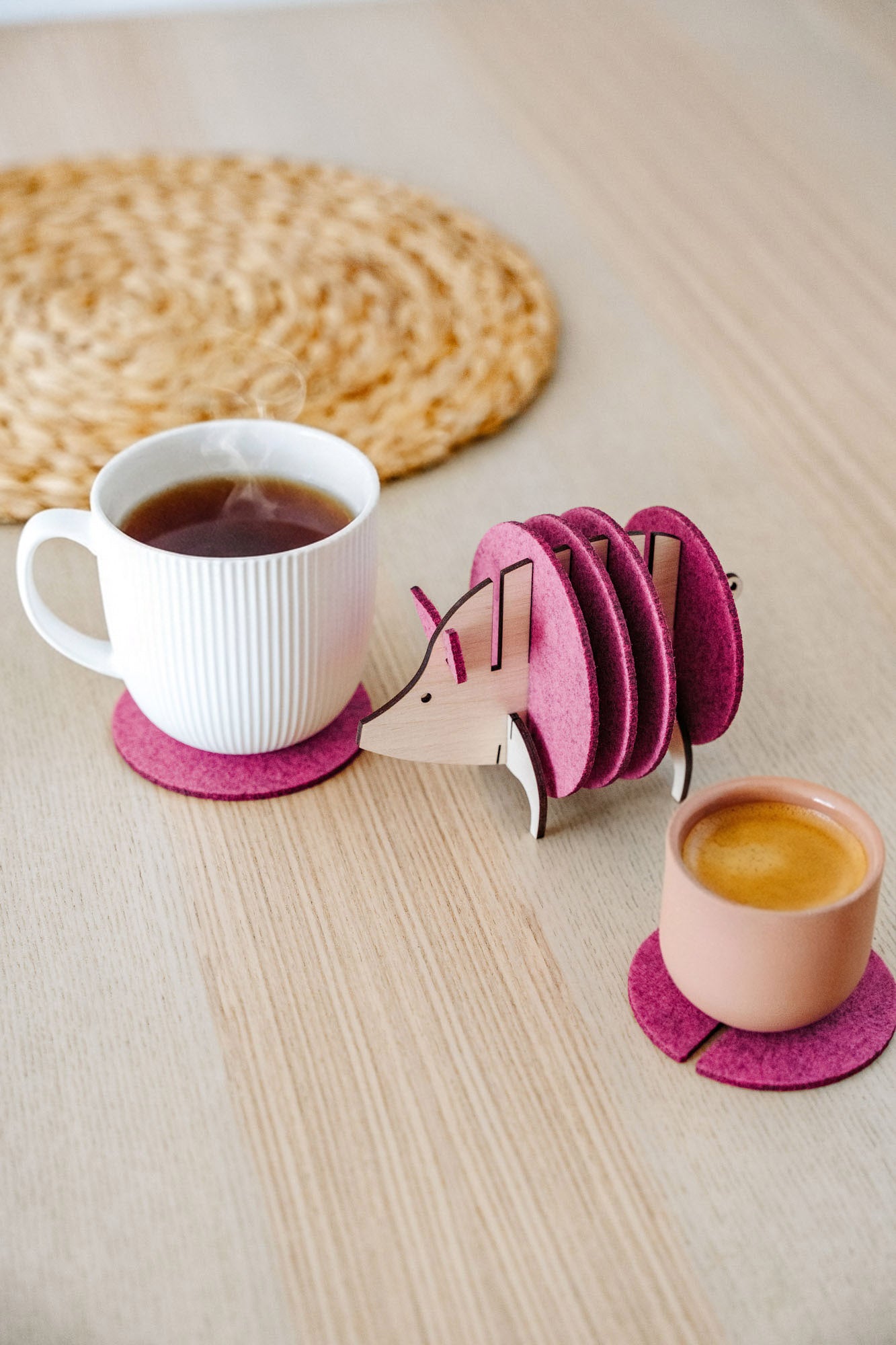 A white cup and a pink cup sit on purple felt coasters next to a wooden hedgehog-shaped coaster holder, with more felt coasters inside it, on a light wooden table—a lovely housewarming gift. A woven mat is in the background.