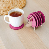 A white ribbed mug on a pink felt coaster sits next to a pink, hedgehog-shaped coaster holder on a light wooden table. A woven placemat is partially visible—perfect as a charming housewarming gift.