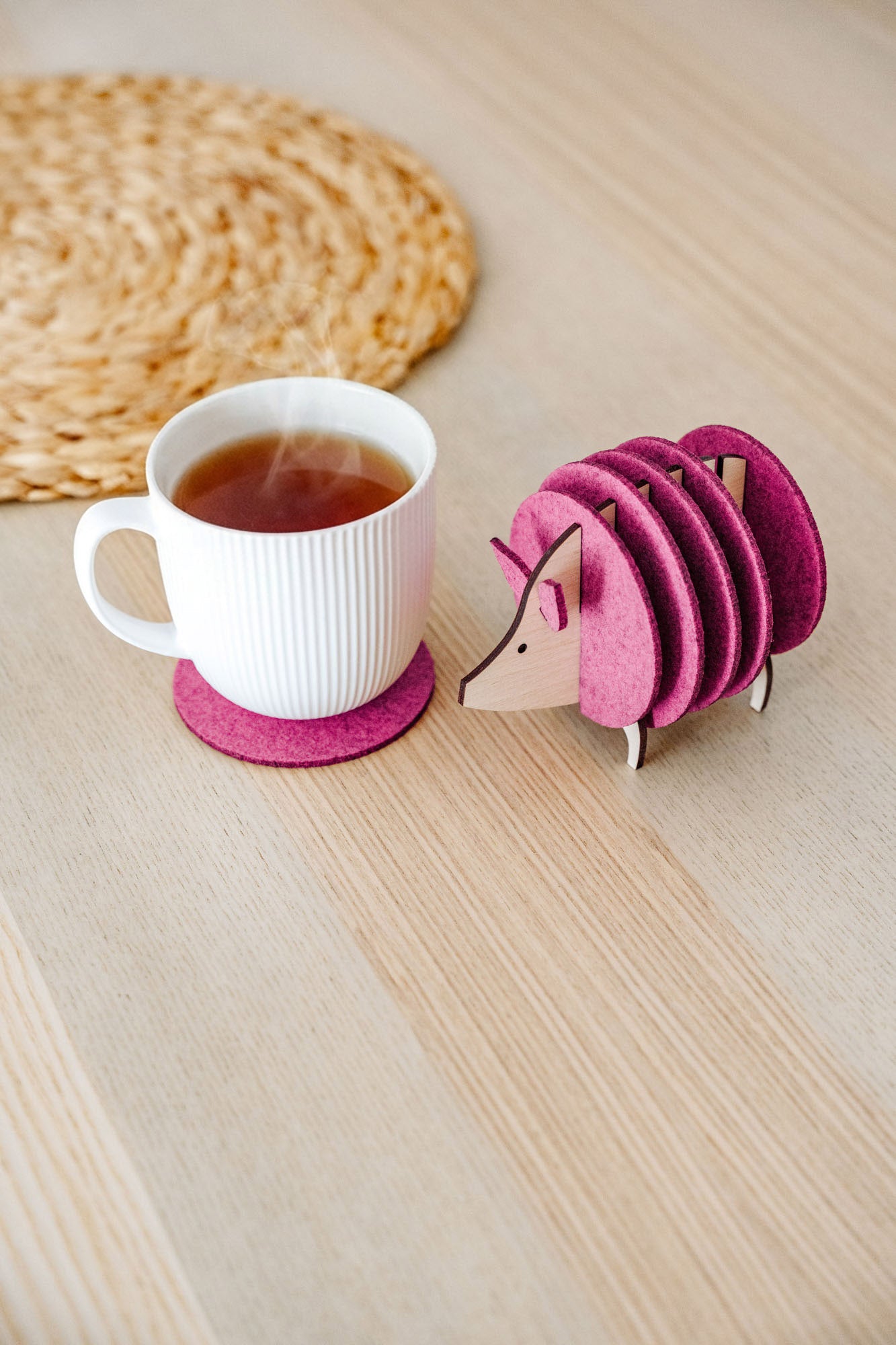A white ribbed mug on a pink felt coaster sits next to a pink, hedgehog-shaped coaster holder on a light wooden table. A woven placemat is partially visible—perfect as a charming housewarming gift.