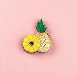 Two cookies on a pink background: one is handcrafted and shaped like a yellow pineapple ring, while the other resembles a whole pineapple with green leaves and a textured cream-colored surface—resembling a sweet Pineapple Pin accessory.