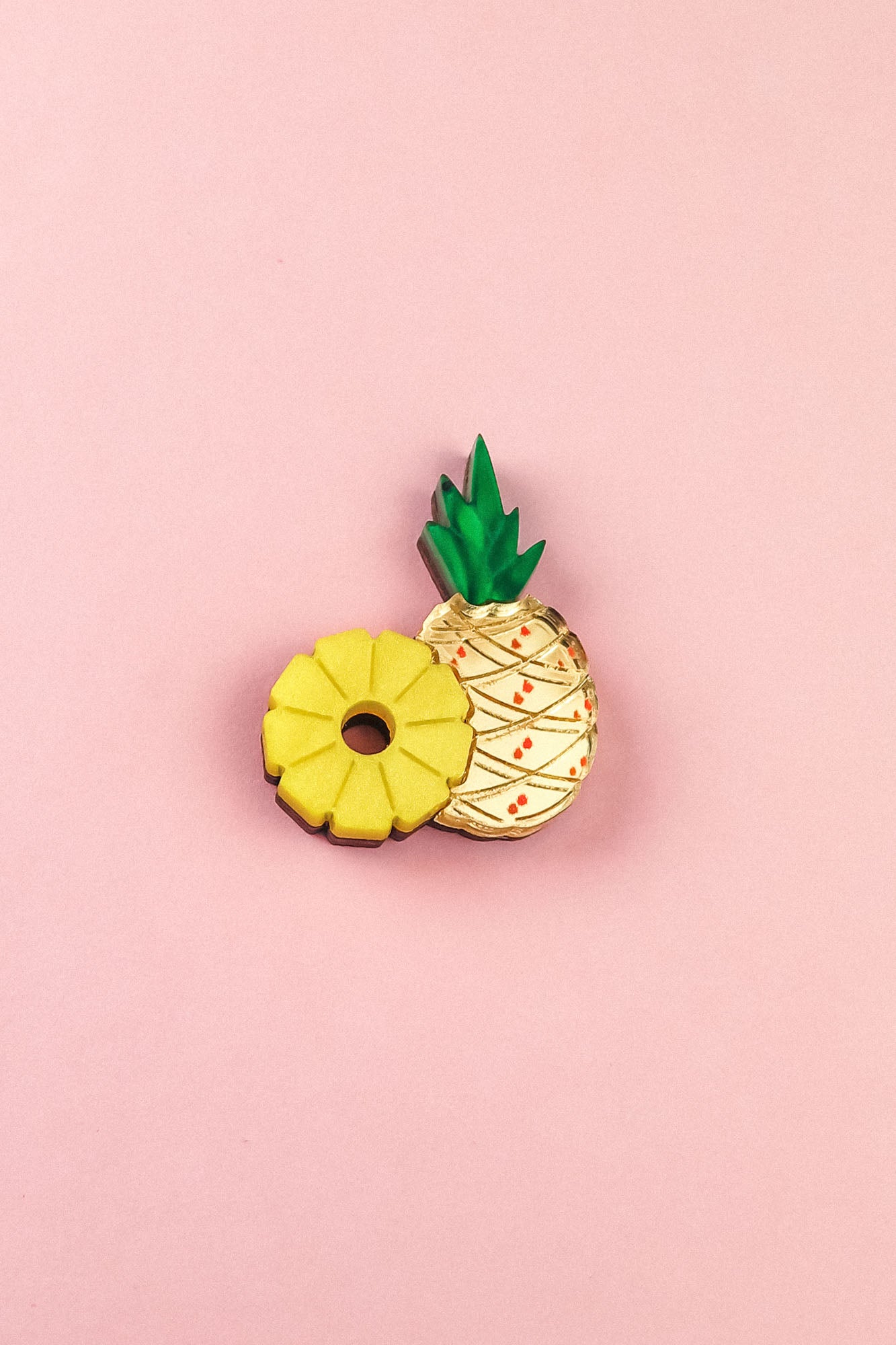 Two cookies on a pink background: one is handcrafted and shaped like a yellow pineapple ring, while the other resembles a whole pineapple with green leaves and a textured cream-colored surface—resembling a sweet Pineapple Pin accessory.