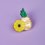 A handcrafted Pineapple Pin with green leaves and gold detailing stands on a light purple background, next to a yellow ring-shaped accessory resembling a sliced pineapple.